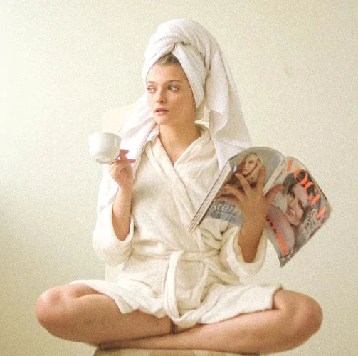 Image of a woman indulging in self-care, relaxing in a white bathrobe while reading a magazine, and sipping tea, embodying the essence of a peaceful and rejuvenating spa experience