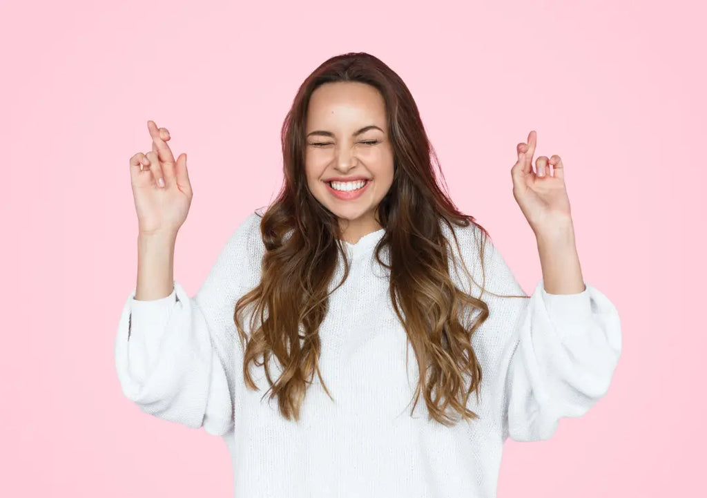 tan woman crossing her fingers in front of a pink background