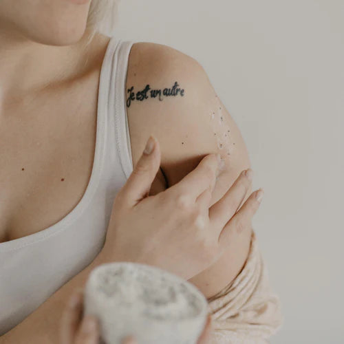 Image of a woman with a tattoo on her shoulder, wearing a white tank top and holding her arm, showcasing her unique style and body art