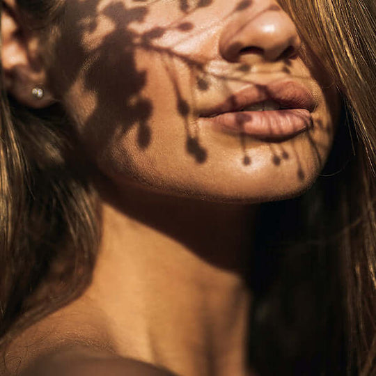 Image of tanned woman with bronzed skin, standing in front of a vibrant sunset. The warm light illuminates her face, and a delicate shadow of a plant falls over her cheek. The woman has a serene expression, and her hair blows gently in the breeze.